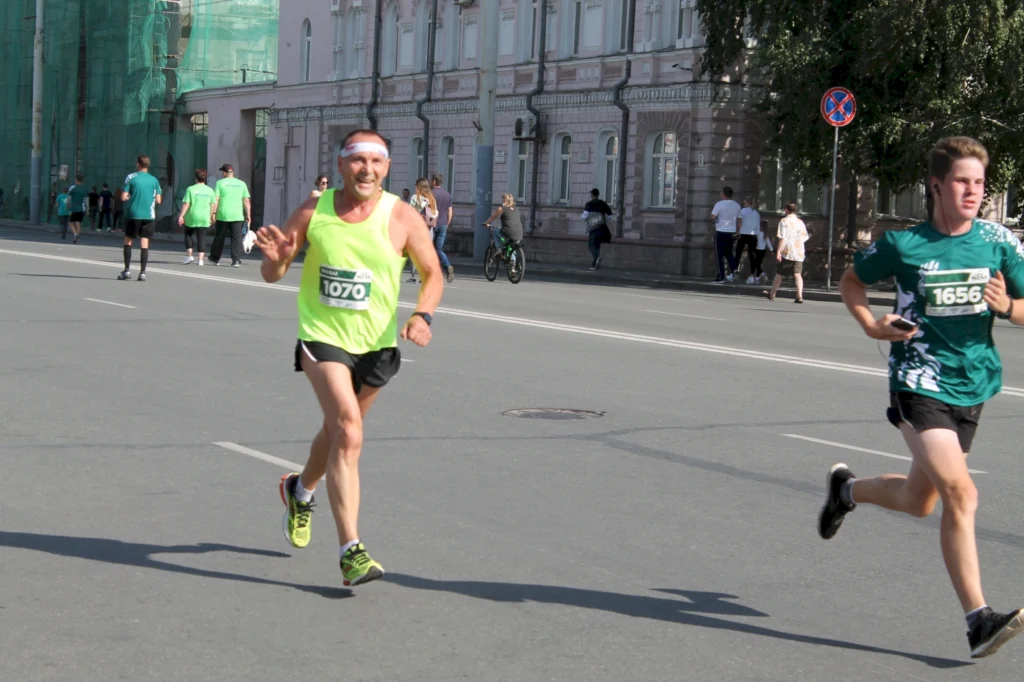 Läufer in lebhaftem grünen Outfit beim Straßenrennen, mit Nummern auf der Brust, während er auf einer urbanen Straße läuft. Im Hintergrund sind weitere Teilnehmer und Zuschauer zu sehen.