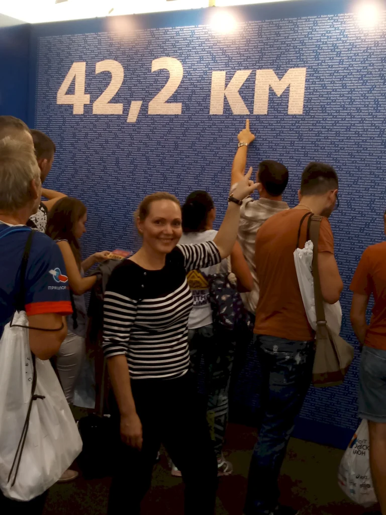 Eine Gruppe von Menschen steht vor einer Wand mit der Aufschrift "42,2 KM". Eine Frau in gestreiftem Oberteil lächelt und zeigt auf die Zahl. Die Umgebung ist lebhaft und zeigt Menschen, die sich für das Event interessieren.