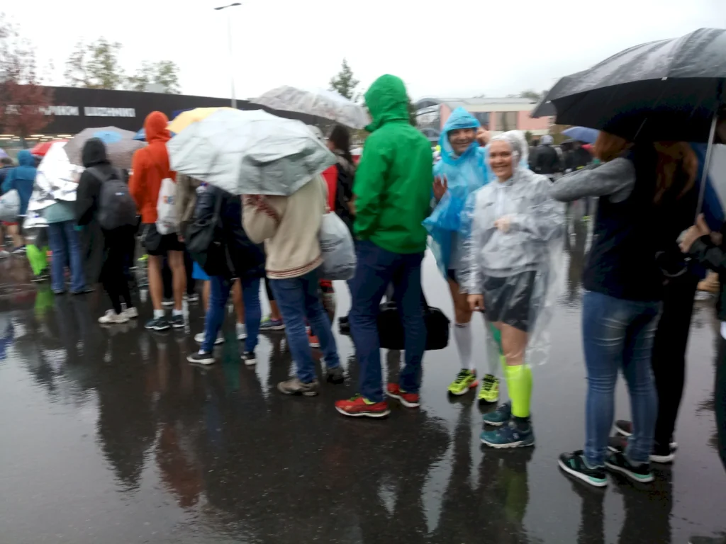 Menschen stehen in einer langen Schlange bei Regenwetter, während sie Regenschirme und Regenjacken tragen. Die Szene zeigt Teilnehmer eines Events oder Wettkampfs, die geduldig auf ihren Eintritt warten. Der Boden ist nass und spiegelt die Umgebung wider.