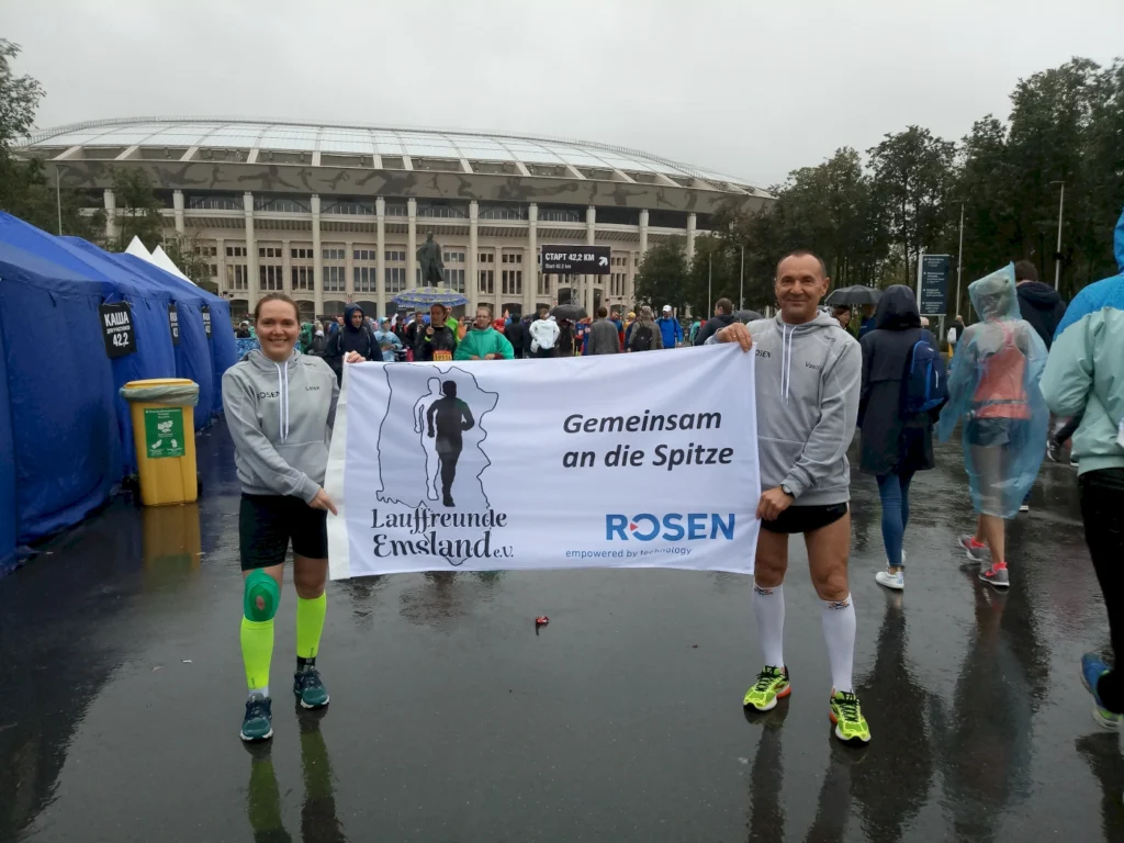 Teilnehmer des Lauf-Events in Emsland halten ein Banner mit der Aufschrift "Gemeinsam an die Spitze" vor einem Stadion, während es regnet. Die Szene zeigt die Vorfreude und den Teamgeist der Läufer.
