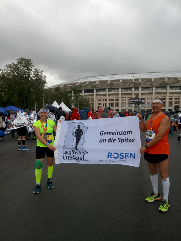 Zwei Läufer halten ein Banner mit der Aufschrift "Gemeinsam an die Spitze" während eines Marathons vor dem Stadion. Im Hintergrund sind zahlreiche Teilnehmer und ein bewölkter Himmel zu sehen.