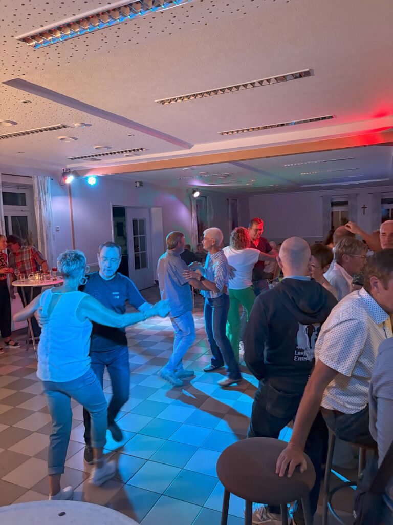 Personen tanzen bei einer Party in einem Raum mit bunter Beleuchtung und entspannter Atmosphäre.