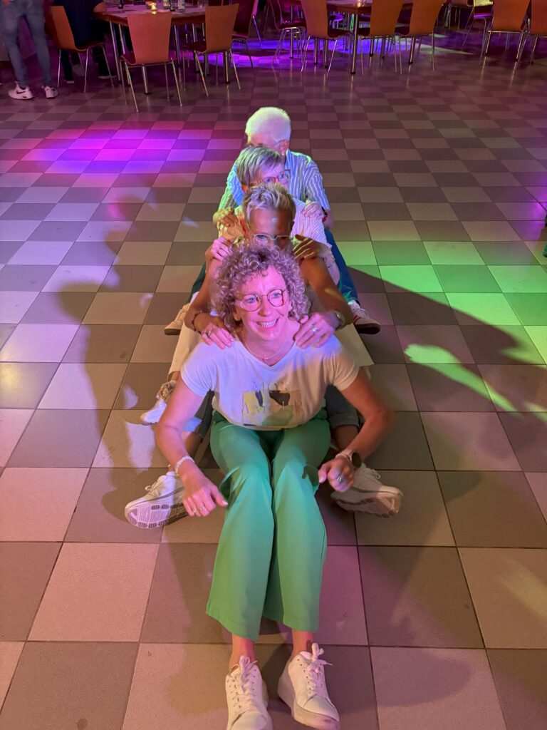 Gruppe von vier Personen tanzt in einer Reihe auf einer beleuchteten Tanzfläche in einem Saal mit bunten Lichtern.