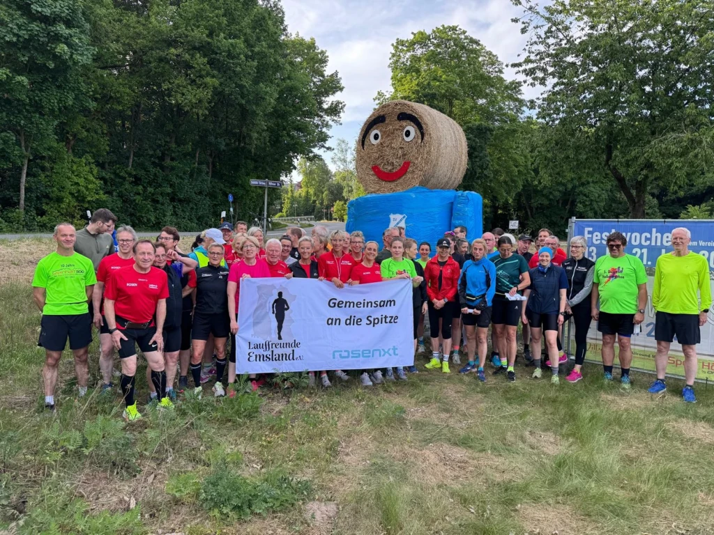 Gruppe von Läufern mit Banner „Gemeinsam an die Spitze“ vor großer Strohfigur, umgeben von grünen Bäumen.