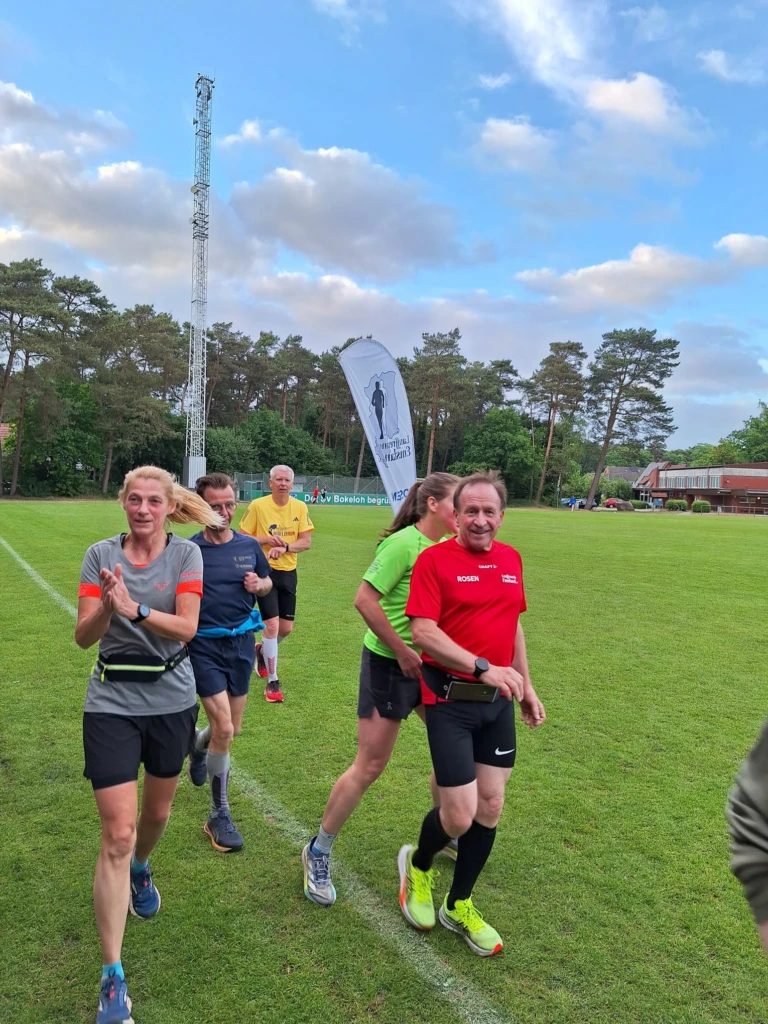 Läufergruppe beim Sportevent auf grüner Wiese mit Wald und Himmel im Hintergrund, Banner im Bild.