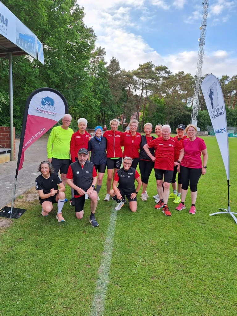 Gruppe von LäuferInnen in roter Sportkleidung auf grünem Rasen bei einer Veranstaltung, zwei Werbebanner im Hintergrund.