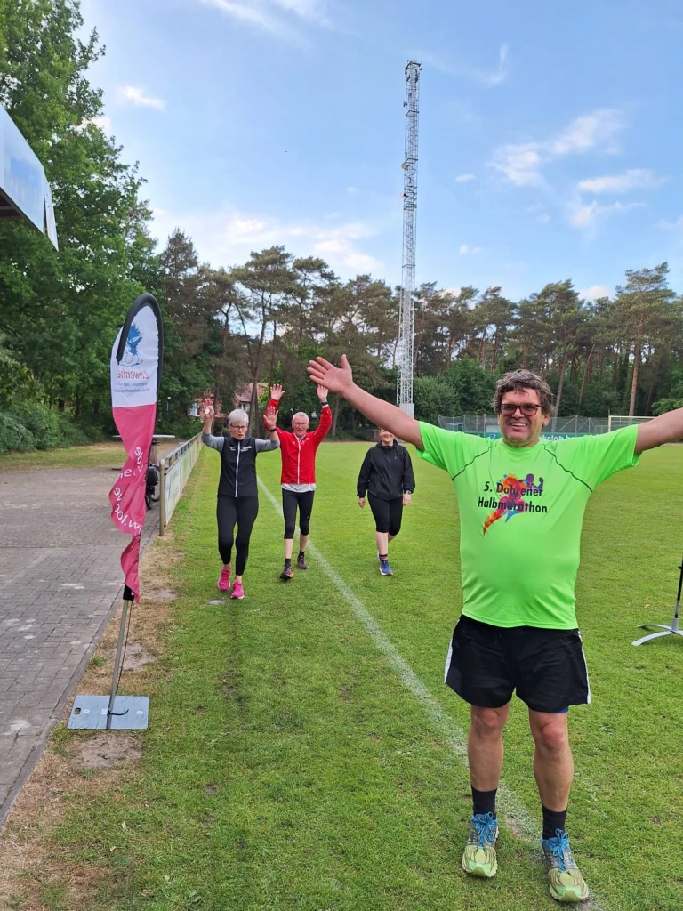 Teilnehmer des Halbmarathons jubeln beim Zieleinlauf auf dem Sportplatz. Bunte Kleidung, Freude und Stolz erkennbar.