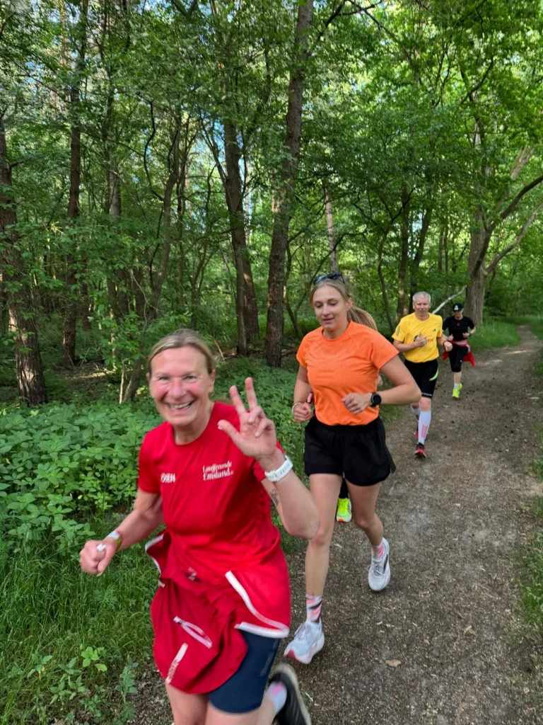 Laufgruppe joggt fröhlich durch grünen Waldweg, Frau vorn in rotem Shirt zeigt „Victory“-Zeichen.