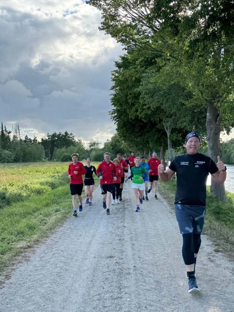 Läufergruppe auf einem Waldweg bei bewölktem Himmel, vorderer Mann hebt Daumen hoch.