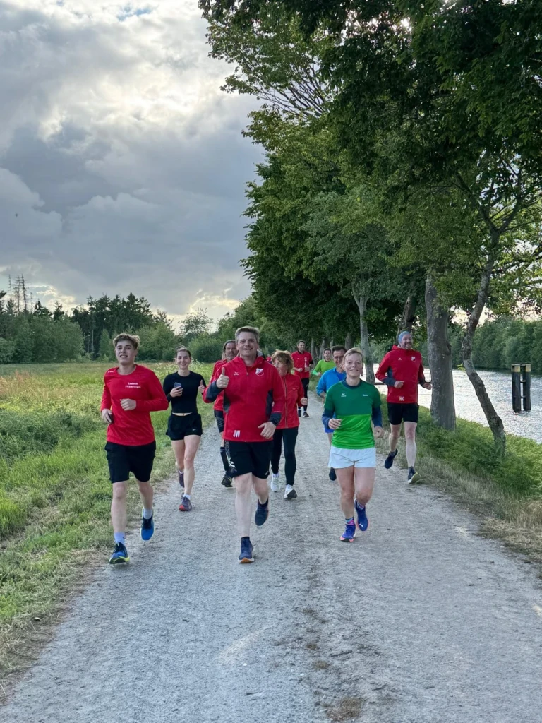 Läufergruppe im Freien auf Naturweg bei bewölktem Himmel, umgeben von Bäumen und einem Fluss.