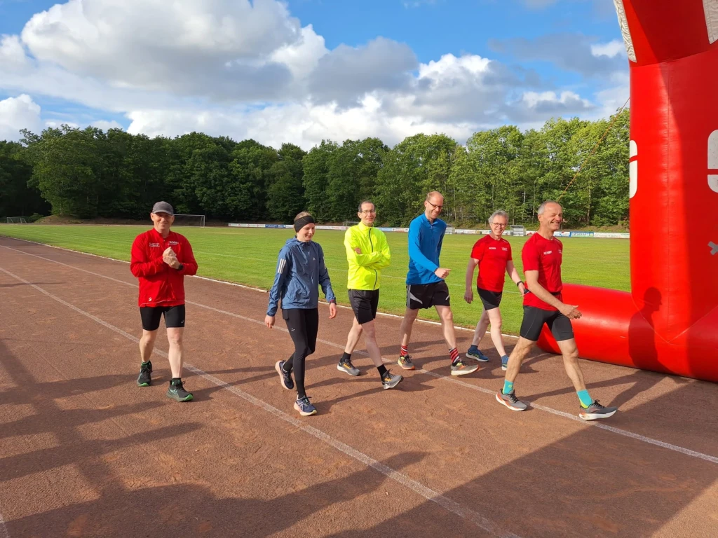 Gruppe von sechs Menschen in Sportkleidung geht über eine Laufbahn im Freien an einem sonnigen Tag.