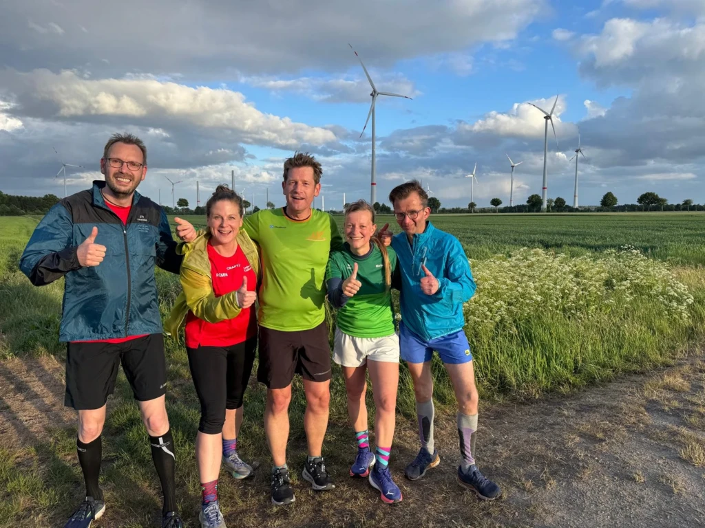 Gruppe von fünf Läufer:innen lächelt mit Daumen hoch vor Windrädern und Feldern im Sonnenschein. Outdoor-Sport, Natur.