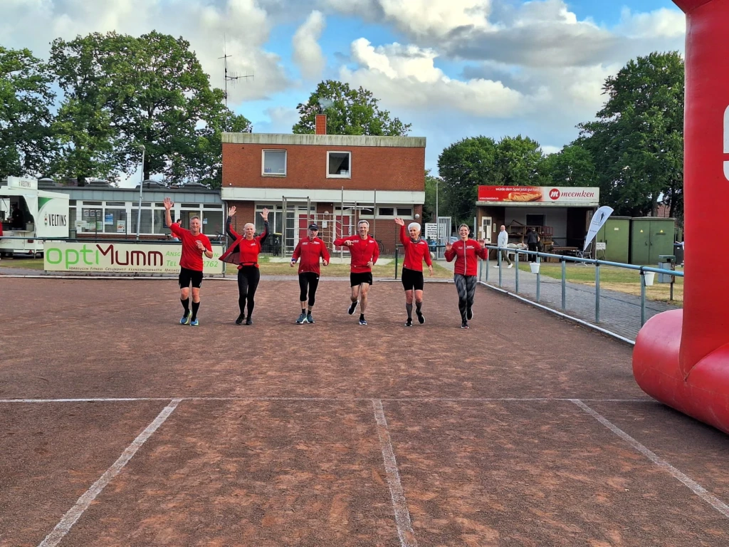 Gruppe von sechs Läufern in roten Trikots jubelt beim Zieleinlauf auf einer Bahn vor einem Vereinsgebäude.