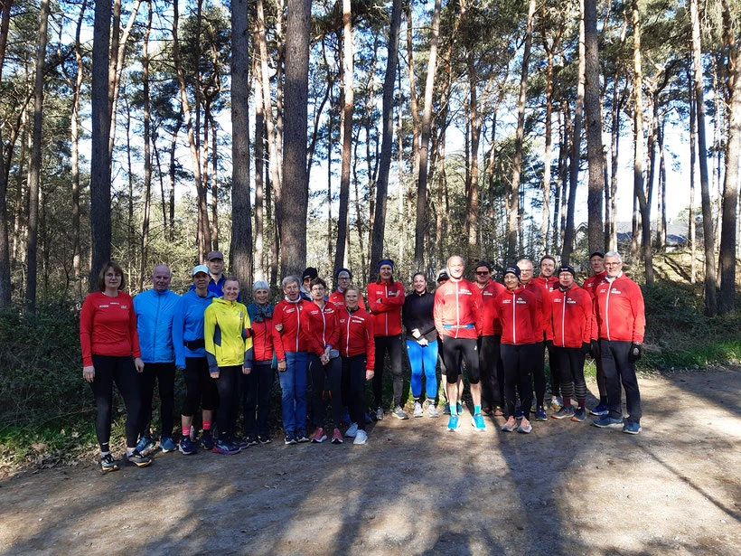 Gruppe von Läufern im Wald, alle in bunter Sportkleidung, lächeln in die Kamera an einem sonnigen Tag.