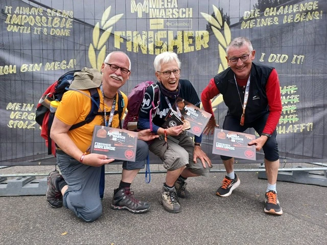 Drei Personen feiern ihren Erfolg beim MegaMarsch mit Finisher-Urkunden vor einer Bannerwand.